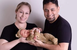 Mujer rubia y hombre con barba sostienen a un bebé en traje de oso, todos sonriendo en un fondo claro.