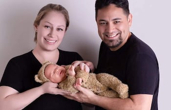 Mujer rubia y hombre con barba sostienen a un bebé en traje de oso, todos sonriendo en un fondo claro.
