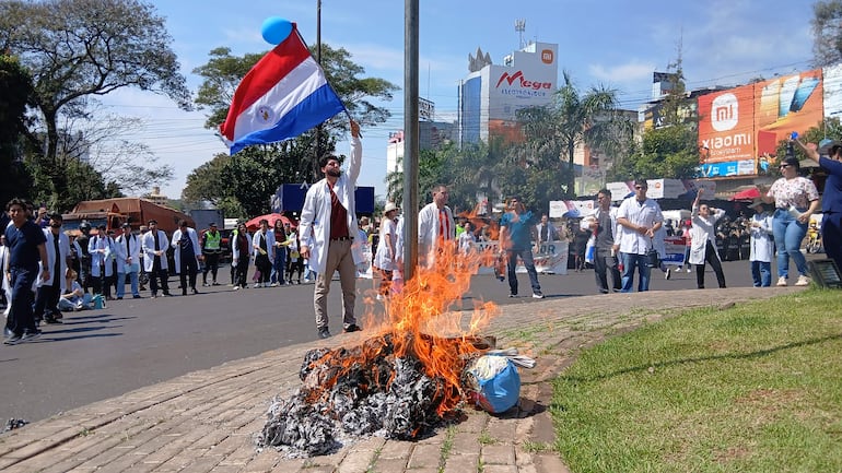 Médicos en batas blancas participan en protesta, uno sostiene una bandera, hay un fuego en la calle.