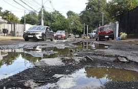 Calle de Santo Domingo con vehículos rojo y negro transitan lentamente entre baches y charcos de agua en un entorno residencial.