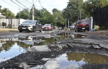 Calle de Santo Domingo con vehículos rojo y negro transitan lentamente entre baches y charcos de agua en un entorno residencial.