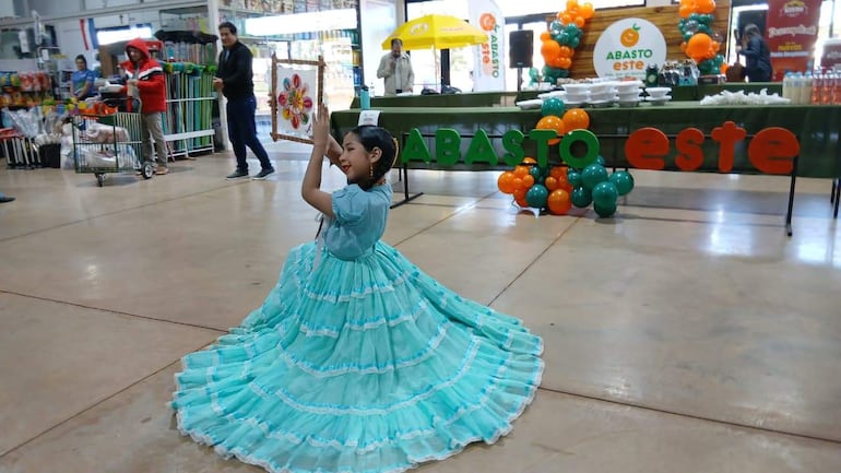Niña en vestido azul con volantes gira mientras sostiene un cuadro decorativo, rodeada de decoraciones de globos verdes y naranjas.