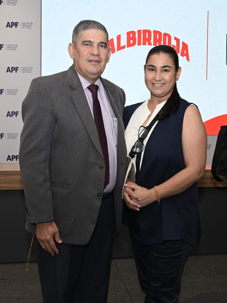 Hombre de traje gris y corbata sonríe junto a mujer con blusa oscura en ambiente formal, fondo con inscripciones de la APF y Albirroja.