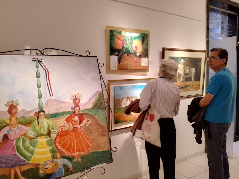 Mujer canosa en vestido claro y hombre con gafas observan pinturas vibrantes en una galería de arte.