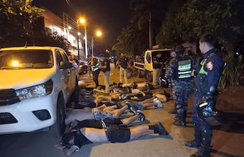 Hombres boca abajo en la calle, varios policías en pie supervisando la detención, ambiente urbano y vehículos de policía presentes.