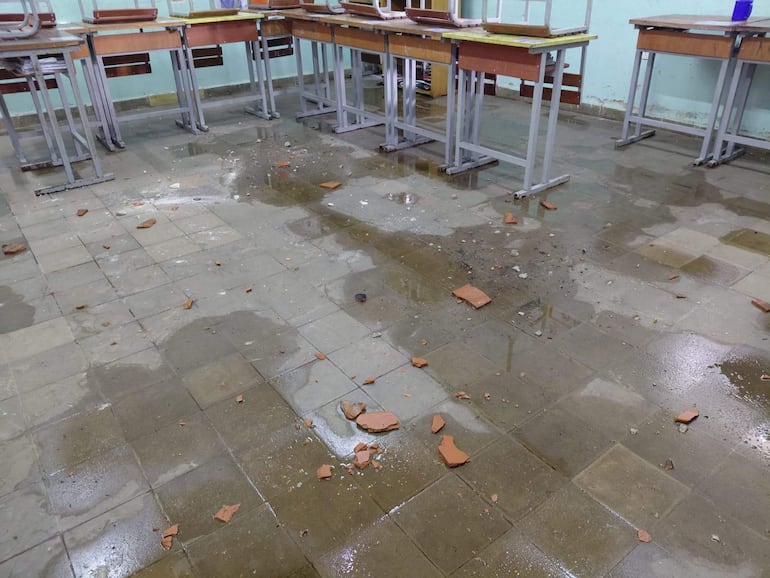 Aula con piso mojado y escombros, mesas y sillas desordenadas tras el temporal, sin personas presentes.