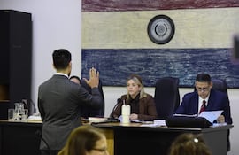 Hombre de espaldas en traje gris levanta la mano, mientras dos personas lo observan en una sala de tribunal con mural de la bandera paraguaya.