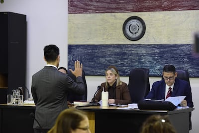 Hombre de espaldas en traje gris levanta la mano, mientras dos personas lo observan en una sala de tribunal con mural de la bandera paraguaya.