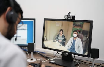 Mujer con auriculares enfocada en una computadora, que muestra a un médico con mascarilla y paciente en consulta virtual.
