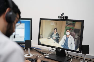 Mujer con auriculares enfocada en una computadora, que muestra a un médico con mascarilla y paciente en consulta virtual.