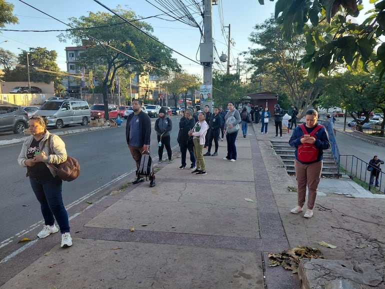 Varias personas en filas, algunas mirando sus teléfonos, en un entorno urbano frente a edificios y coches.