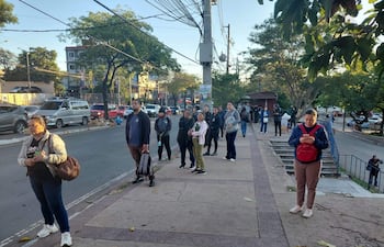 Varias personas en filas, algunas mirando sus teléfonos, en un entorno urbano frente a edificios y coches.
