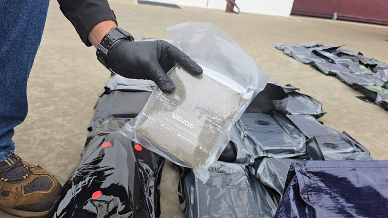 Persona con guantes sosteniendo un paquete dentro de un entorno exterior, rodeada de varios paquetes negros en el suelo.
