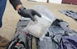Persona con guantes sosteniendo un paquete dentro de un entorno exterior, rodeada de varios paquetes negros en el suelo.