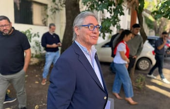 César Ruíz Díaz con traje oscuro y gafas, rodeado de personas en un entorno exterior con vegetación y vehículos.