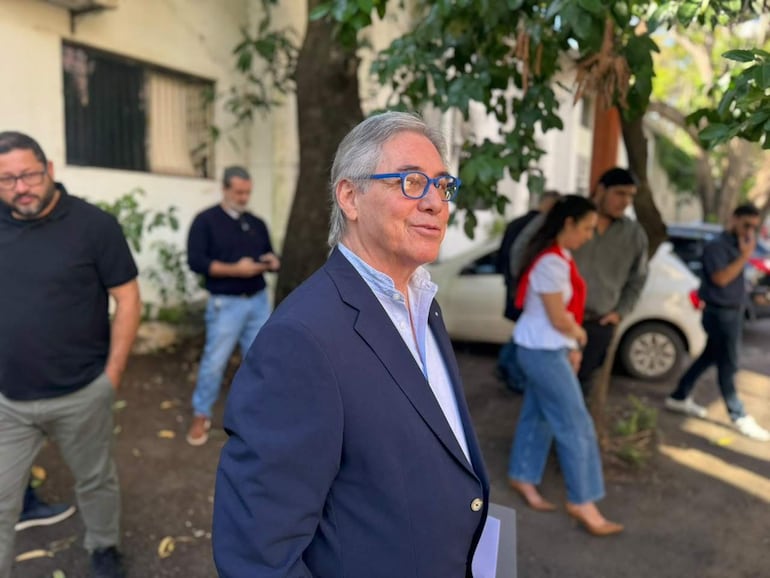 César Ruíz Díaz con traje oscuro y gafas, rodeado de personas en un entorno exterior con vegetación y vehículos.