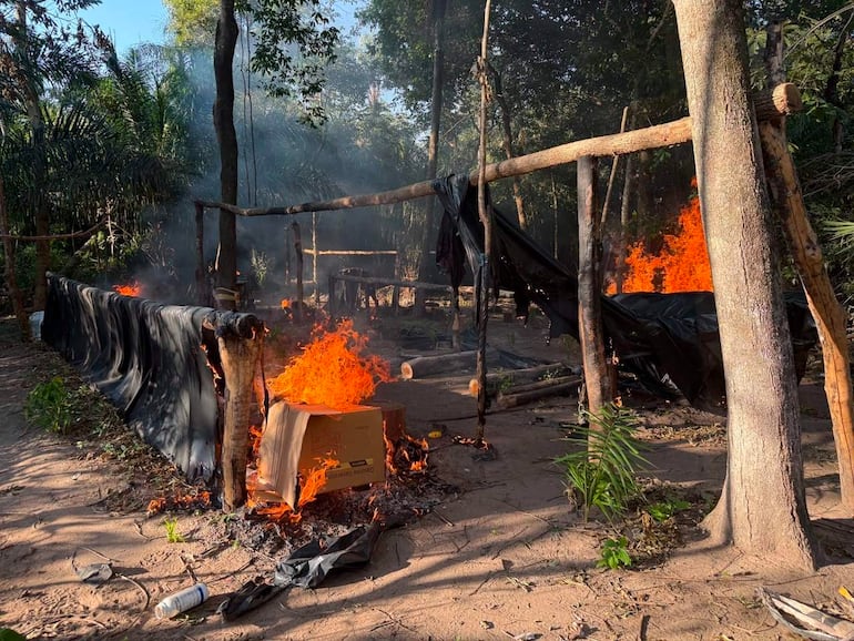 Fuego activo en un campamento improvisado rodeado de vegetación densa y humo ascendiendo al cielo.