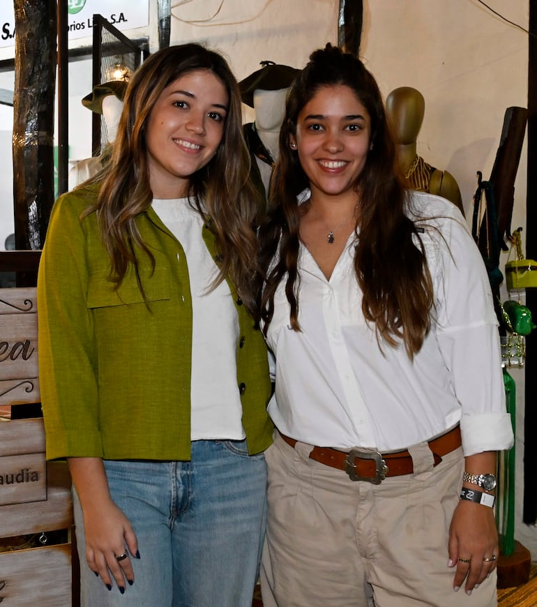 Dos mujeres sonríen, una con chaqueta verde y jeans, otra con camisa blanca y pantalones claros, en un ambiente de mercado.