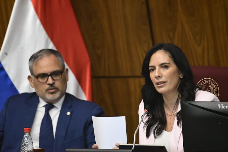 Claudia Centurión habla con papel en mano, vestida con blusa blanca y abrigo rosa, mientras un hombre serio la escucha.