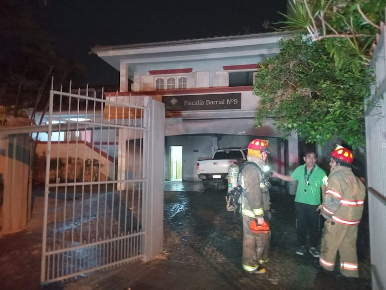 Dos bomberos en trajes protectores conversan con un hombre de camiseta verde frente a la Fiscalía Barrial N°9, con suelo mojado.