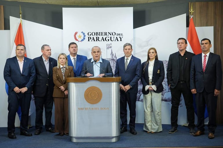 Núñez en chaqueta gris dirige la conferencia, rodeado por siete hombres y una mujer, todos en trajes formales, en un ambiente oficial.