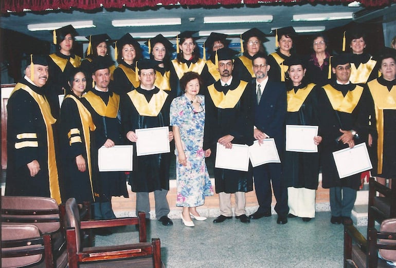 Grupo de 17 graduados en togas negras y birretes amarillos, todos sosteniendo certificados, con una mujer de vestido florido en el centro.