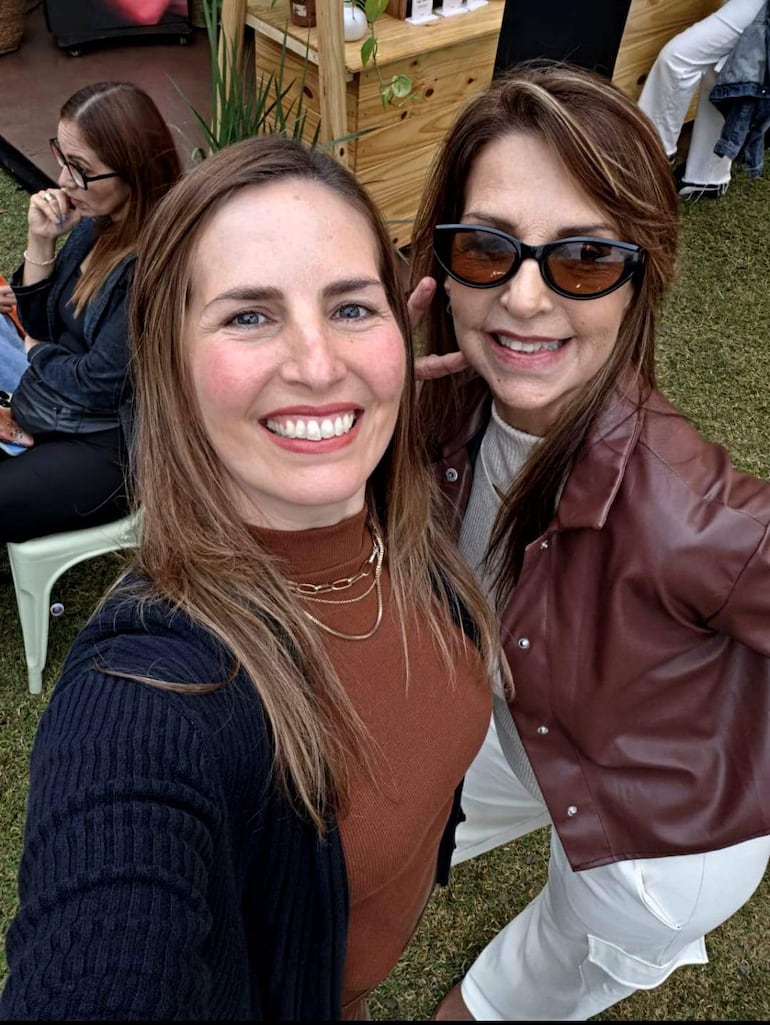 Dos mujeres sonrientes, una con suéter marrón y blusa blanca, y otra con gafas de sol y chaqueta marrón, en un jardín.