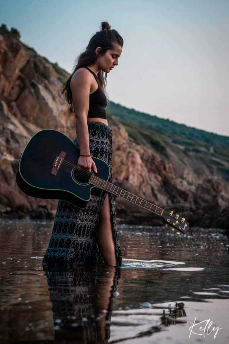 Kelly Ou Moi de perfil, vestida con camiseta negra y falda étnica, sostiene guitarra acústica en entorno natural junto al agua.