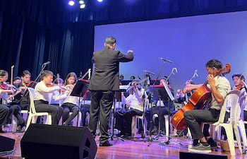 Músicos de la orquesta en sillas blancas, vistiendo camisas blancas, bajo luces azules y púrpuras, mientras el director marca el ritmo.
