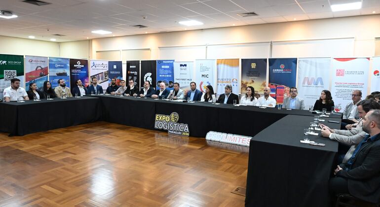 Hombre en traje hablando ante audiencia formal en un salón con banners de empresas de logística.