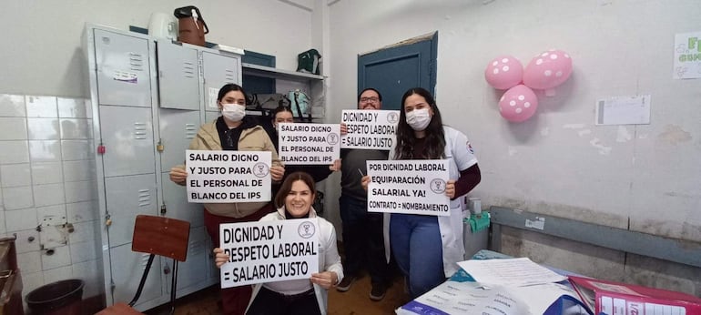 Mujer sentada en silla con cartel que dice 'DIGNIDAD Y RESPETO LABORAL = SALARIO JUSTO', rodeada de cuatro profesionales con mascarillas y carteles similares.