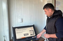 Hombre de perfil introduce un documento en máquina de votación, en un ambiente cerrado con instrucciones en pantalla.