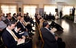 Hombre al micrófono presenta en foro, con audiencia formalmente vestida tomando notas en ambiente de auditorio decorado con plantas.