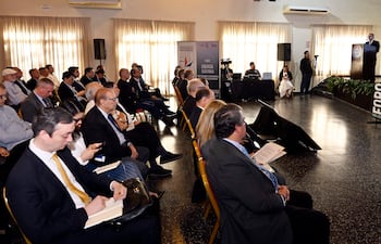 Hombre al micrófono presenta en foro, con audiencia formalmente vestida tomando notas en ambiente de auditorio decorado con plantas.