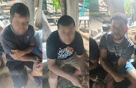 Tres hombres esposados, sentados en un entorno al aire libre con expresión seria y manos cruzadas, rodeados de árboles y mobiliario informal.