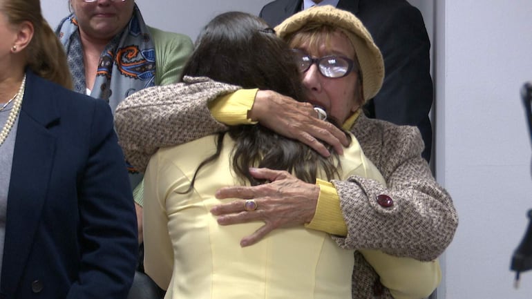 Tania Irún viste blusa amarilla y abraza a Margarita Irún, que usa abrigo marrón y sombrero claro, en un ambiente íntimo.
