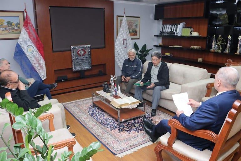 Emiliano Rolón Fernández en reunión formal, sosteniendo un documento, rodeado de cuatro hombres atentos en un ambiente institucional.