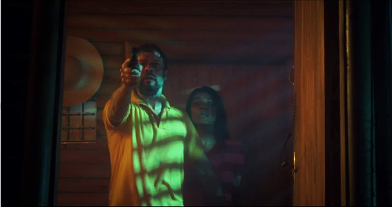 Rafael Alfaro destaca con camiseta amarilla, sosteniendo un objeto, mientras Majo Cabrera, asustada, mira hacia adelante en habitación oscura.