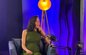 Fiorella Migliore sentada en un sofá azul, con vestido verde y gesticulando, en un ambiente de iluminación suave.