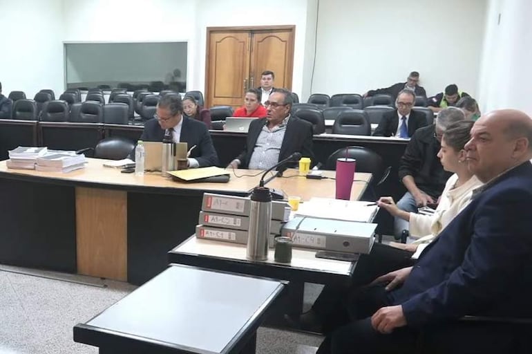 Miguel Robles y otros hombres en traje sentados alrededor de una mesa, en un ambiente formal de tribunal, con documentos y dispositivos electrónicos.