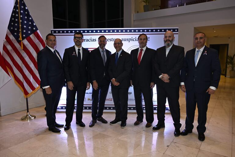 Grupo de seis hombres en trajes formales sonríen ante panel iluminado que agradece a los auspiciantes, con bandera de EE.UU. al fondo.