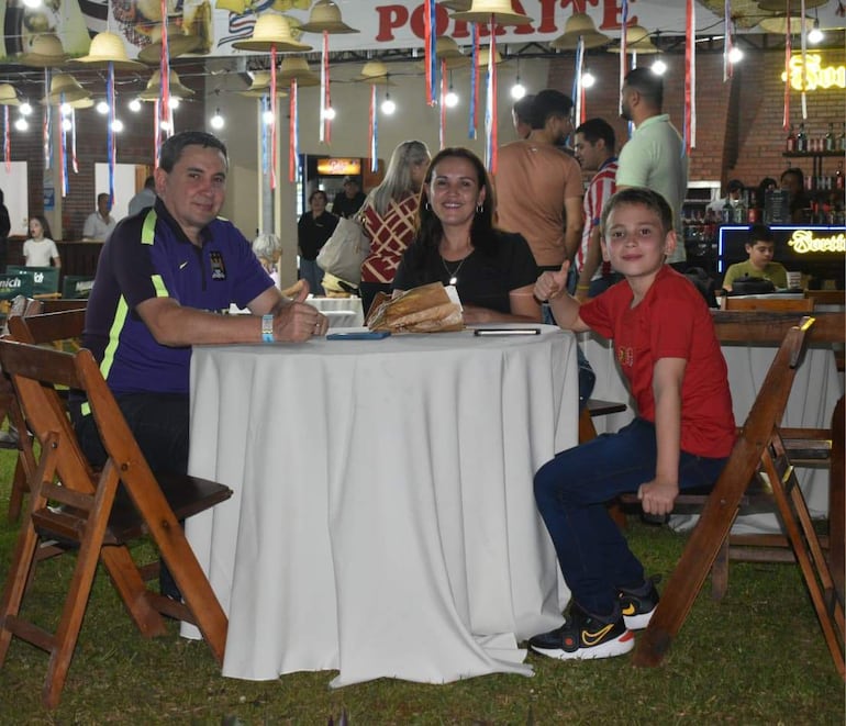 Hombre en camiseta morada, mujer en atuendo negro y niño en camiseta roja, sonriendo en un ambiente festivo de restaurante.