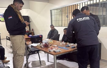 Policía antinarcóticos en ambiente austero con hombres detenidos, inspectando objetos sobre la mesa.