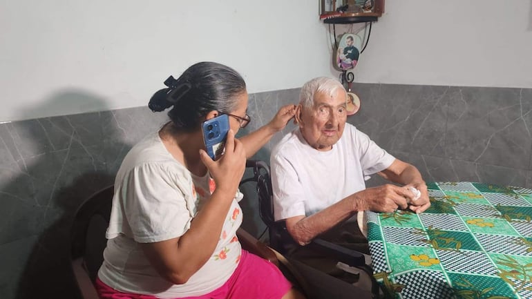 Canuto González, de cabello canoso y en silla de ruedas, con expresión neutral, mientras una mujer de cabello recogido habla por teléfono a su lado.