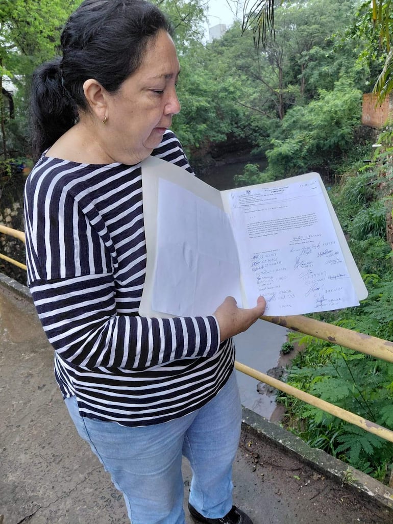Mujer con camiseta a rayas blancas y negras y jeans claros, sostiene hojas mientras examina el entorno natural junto a un camino.