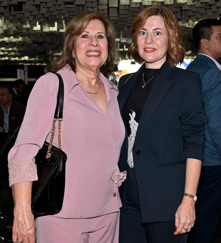Dos mujeres sonríen, una con blusa rosa y cabello rizado, otra en traje negro y corte bob, en un ambiente moderno.