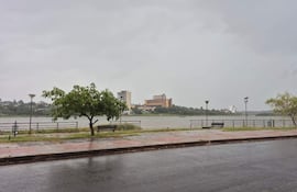 Paisaje urbano costero con río Paraná de fondo, edificios, árboles y vereda vacía en un día gris y lluvioso.