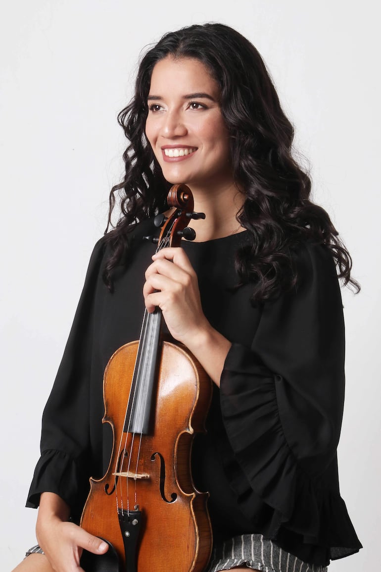 Mujer con cabello rizado y blusa negra sonríe, sosteniendo un violín en un fondo blanco.