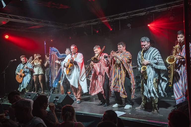 Ocho músicos con ponchos coloridos tocan en un escenario oscuro, iluminado por luces rojas, frente a un público entusiasta.