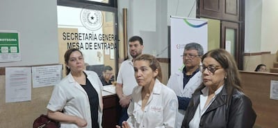 Isabel Morán y otra mujer en bata blanca, con expresión preocupada, conversan en oficina con hombres al fondo, todos serios.